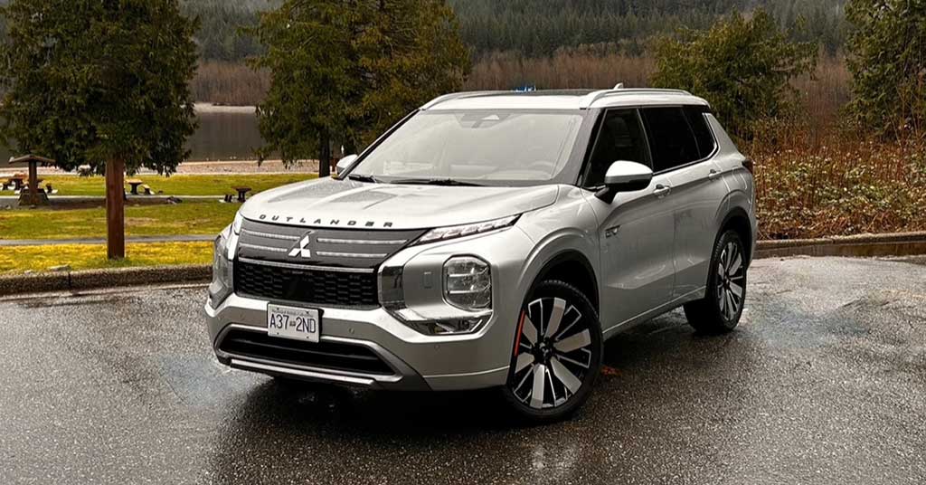 2026-Outlander-PHEV - Strathmore Voice Front-three-quarter view of a silver Mitsubishi Outlander SUV parked on a wet pavement with trees and a lake in the background, daytime outdoor scene.