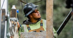 Construction worker wearing a hard hat, safety glasses, and overalls holds a perforated metal panel while looking upward on a construction site.